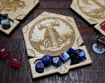 May include: A wooden dice tower with a laser-engraved image of a giant hammer and a mountain range. The tower has a compartment for dice, and there are several blue dice inside the compartment.
