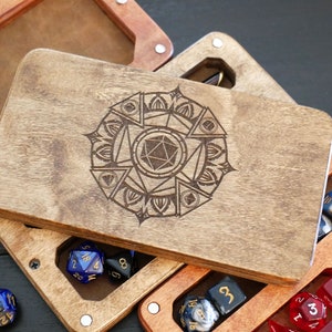 May include: A wooden box with a lid that opens to reveal a compartment for dice. The lid has a laser-engraved design of a mandala with a d20 in the center. The box is made of light brown wood and has a natural finish.