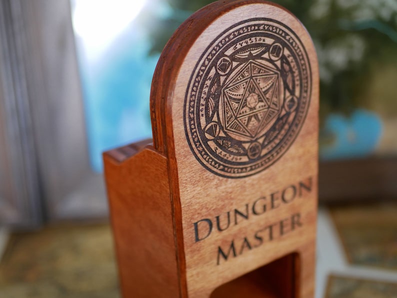 Puede incluir: Una caja de madera con una ranura para dados y las palabras "Dungeon Master" grabadas en la parte delantera. La caja tiene un dise&ntilde;o circular con un dado estilizado en el centro.