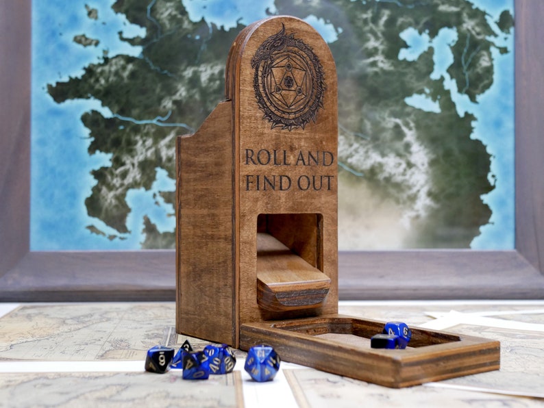 Puede incluir: Una torre de dados de madera con una tapa con bisagras y una bandeja. La torre tiene un dise&ntilde;o geom&eacute;trico y el texto "Roll and Find Out" grabado en la parte delantera. La bandeja est&aacute; abierta y hay varios dados azules y negros dentro.