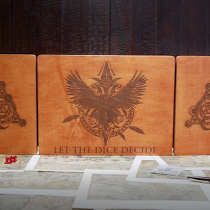 May include: A three-piece wooden box set with a brown leather-like finish. The center panel features a stylized bird with wings spread and the text "Let the Dice Decide". The side panels feature a circular design with a crescent moon and a die.