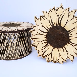 May include: A wooden box with a sunflower design on the lid and sides. The box is made of light wood and has a lattice pattern on the sides. The sunflower design is brown and yellow.