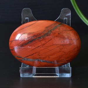 Könnte beinhalten: Ein polierter, oval geformter roter Jaspisstein mit schwarzen und grauen Adern, präsentiert auf einem transparenten Acrylständer. Die Oberfläche des Steins ist glänzend und reflektiert das Licht. Der Jaspis ist ein natürlicher Edelstein.