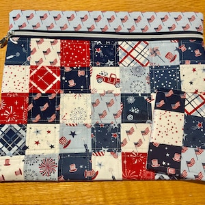May include: A quilted zippered pouch and matching small wallet with a patchwork design in red, white, and blue. The pattern includes American flags, fireworks, and stars. The pouch has a light blue flag-patterned border.
