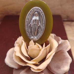 Könnte beinhalten: Eine ovale silberne religiöse Medaille mit einer Darstellung der Jungfrau Maria und dem Text "O Mary Conceived Without Sin Pray For Us Who Have Recourse To Thee 1830" auf einem ovalen grünen Sockel. Die Medaille liegt auf einer künstlichen beige-braunen Blume.