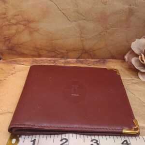 May include: A burgundy leather wallet with gold accents and the Cartier logo embossed on the front. The wallet is measured with a tape measure, showing it is approximately 3 inches wide.