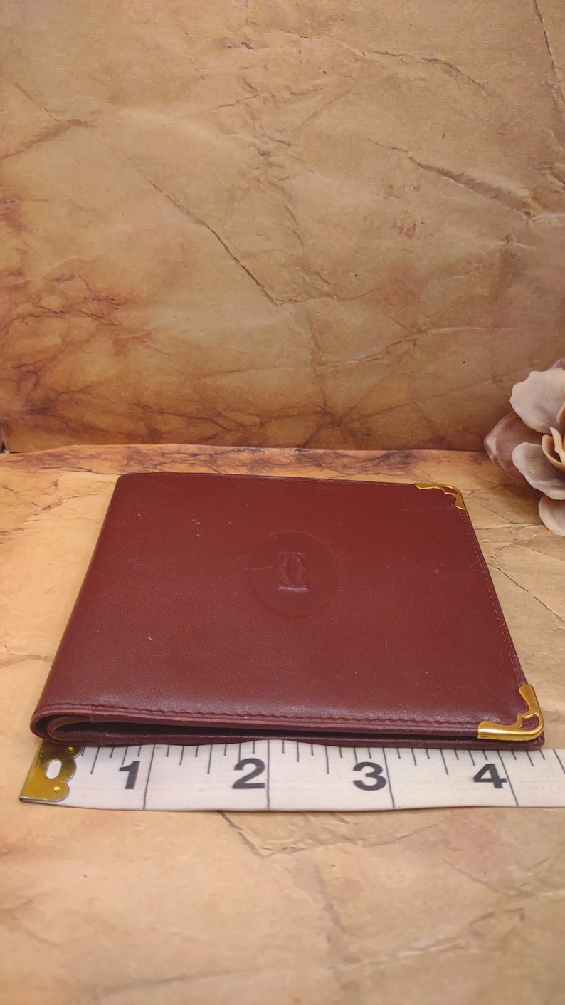 May include: A burgundy leather wallet with gold accents and the Cartier logo embossed on the front. The wallet is measured with a tape measure, showing it is approximately 3 inches wide.