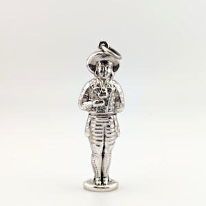 May include: A silver-colored charm in the shape of a person wearing a hat and a detailed outfit. The charm has a loop at the top for attaching to a bracelet or necklace. The figure is standing upright, with intricate detailing on the clothing.