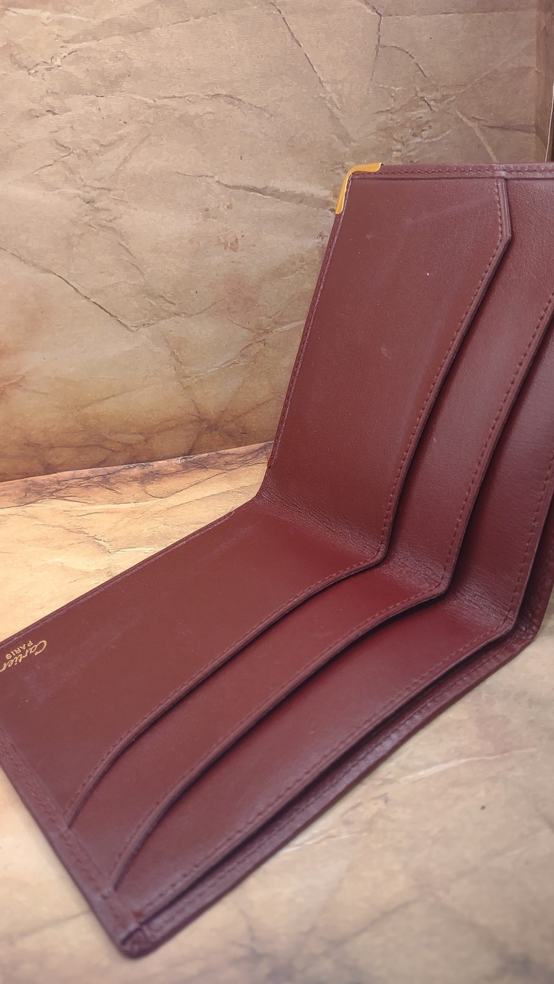 May include: A burgundy leather wallet with gold accents and the text "Cartier Paris" embossed on the front.