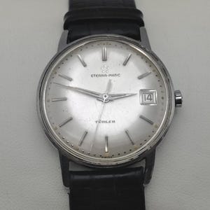 May include: A silver wristwatch with a white face and black leather band. The watch face has black hands, black hour markers, and a date window at the 3 o'clock position. The watch face also has the text "ETERNA-MATIC" and "TÜRLER" printed on it.