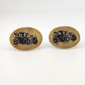 May include: A pair of gold-coloured cufflinks with an oval shape. Each cufflink features a detailed, black and silver vintage car design set against a textured, sunburst-like background.