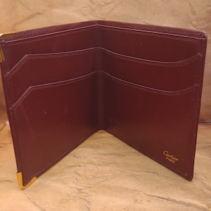 May include: A burgundy leather wallet with gold accents and the text "Cartier Paris" on the inside.