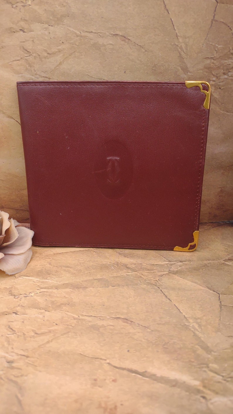 May include: A burgundy leather wallet with gold accents and the Cartier logo embossed on the front.