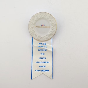 Peut inclure: Une épingle en céramique blanche avec une bordure dorée et le mot "Millennium" imprimé dessus. Un ruban blanc avec une bordure bleue et le texte "Ask me how to become the Lenox Millennium Bride and Groom" est attaché.