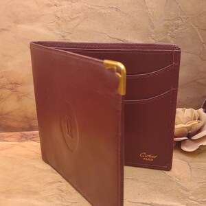 May include: A burgundy leather wallet with gold accents and the Cartier Paris logo. The wallet has multiple card slots and a fold-over closure.
