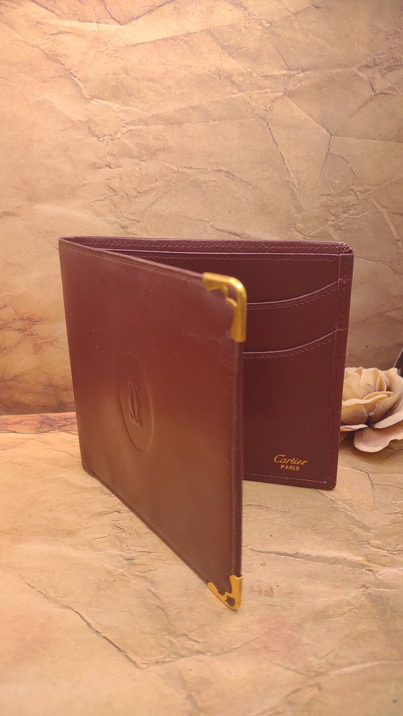 May include: A burgundy leather wallet with gold accents and the Cartier Paris logo. The wallet has multiple card slots and a fold-over closure.