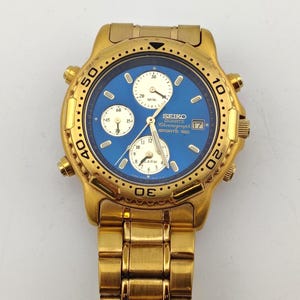 May include: A gold-tone Seiko Quartz Chronograph Sports 150 watch with a blue dial and three cream-colored sub-dials. The watch features a rotating bezel with black numerals, a date window at the three o'clock position, and a metal link band.