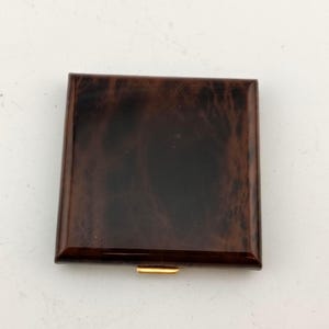 May include: A square, brown, vintage cigarette case with a glossy finish. The case has a marbled pattern and a gold-colored clasp. The case is closed and appears to be made of a hard material.