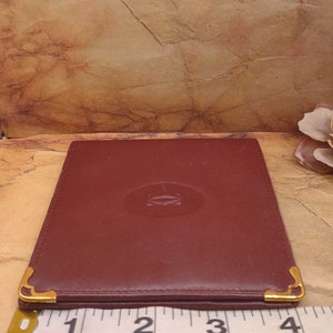 May include: A burgundy leather wallet with gold accents and a gold embossed logo. The wallet is approximately 3 inches wide.