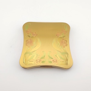 May include: A gold-colored, square compact with rounded edges. The compact features an engraved floral design in shades of peach and green. The floral pattern is symmetrical, with flowers and ribbons. The compact is closed.