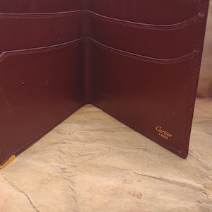 May include: A burgundy leather wallet with multiple card slots and a billfold. The wallet is open and the inside is visible. The brand name "Cartier Paris" is embossed in gold on the inside of the wallet.