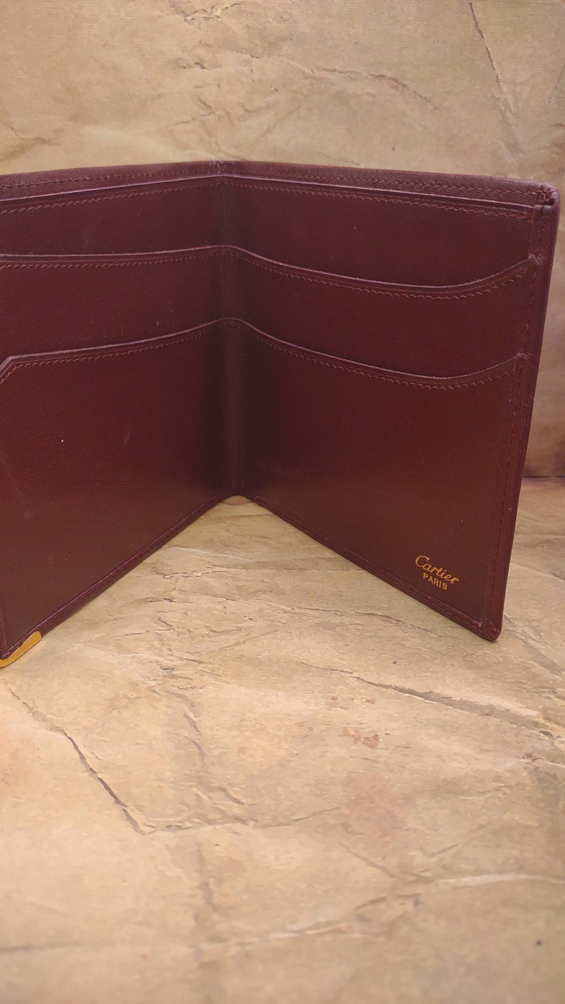 May include: A burgundy leather wallet with multiple card slots and a billfold. The wallet is open and the inside is visible. The brand name "Cartier Paris" is embossed in gold on the inside of the wallet.