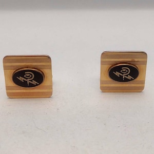 Puede incluir: Un par de gemelos de oro con un diseño de esmalte negro. El diseño presenta una letra "P" estilizada con un rayo.