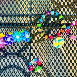 May include: A collection of colorful beaded jewelry, including a bracelet, necklace, and a beaded flower-shaped accessory. The necklace features a lollipop charm and a pink safety pin. The beads are in various colors.
