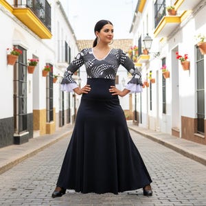 May include: A black and white flamenco dress with a long, flowing skirt. The bodice features a V-neck and a pattern of white polka dots on a black background. The sleeves have ruffled cuffs. The dress is worn in a cobblestone street.