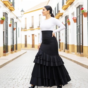 May include: A woman wearing a white long-sleeved top and a high-waisted black skirt with layered ruffles. The skirt has a mermaid silhouette. The woman is standing on a cobblestone street in front of white buildings.