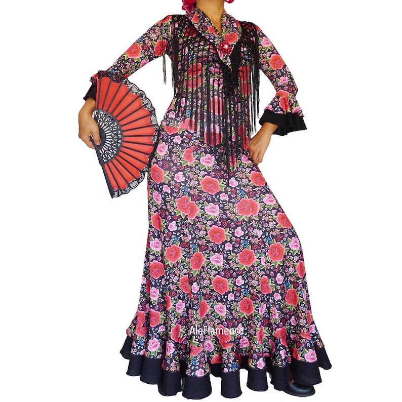 May include: A flamenco dress with a floral pattern of red and pink roses on a black background. The dress has long sleeves with black cuffs, a ruffled hem, and a matching shawl with fringe. A red fan is held in the hand.