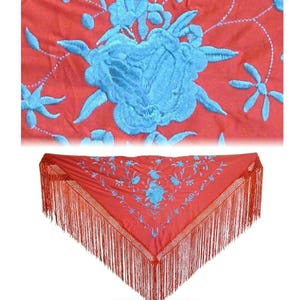 May include: A triangular red shawl featuring intricate light blue floral embroidery across the fabric. The edges of the shawl are finished with long, thin red fringe that hangs down from the bottom and sides of the garment.