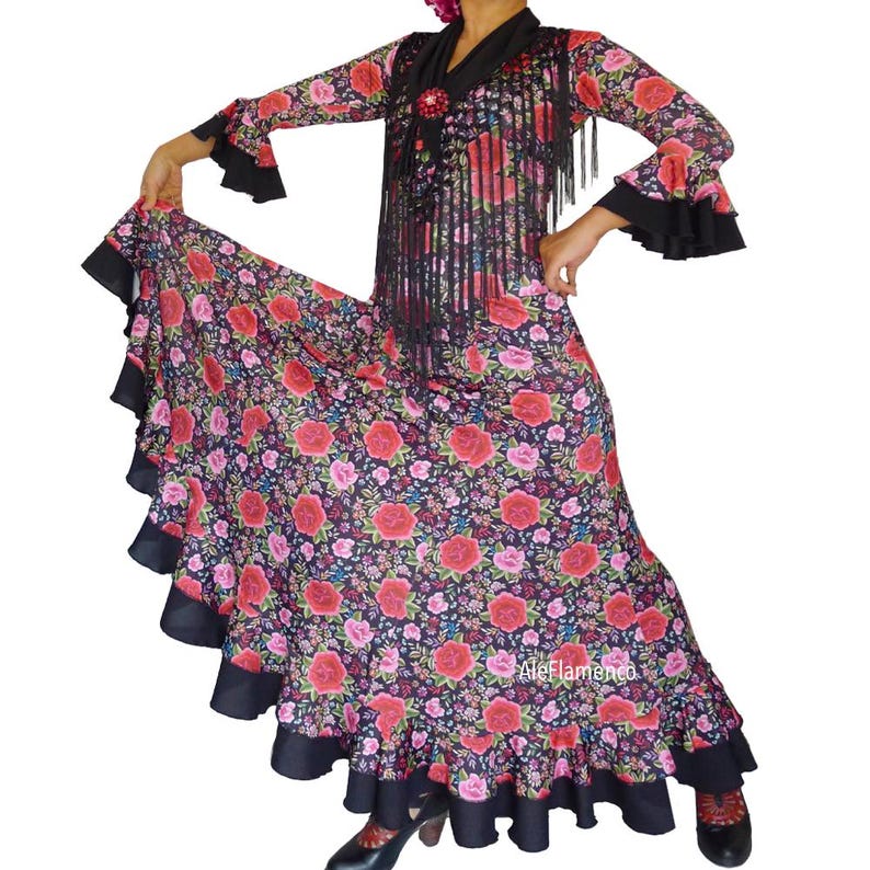 May include: A flamenco dress featuring a floral design of red and pink roses on a dark background. The dress has long sleeves with black ruffled cuffs, a fringed shawl, and a full skirt with a black ruffle at the hem. The dress is designed for dance.