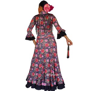 May include: A long-sleeved flamenco dress with a floral pattern of red and pink roses on a dark background. The dress has black fringe and accents, with a matching fan. The dress is designed for dance performances.