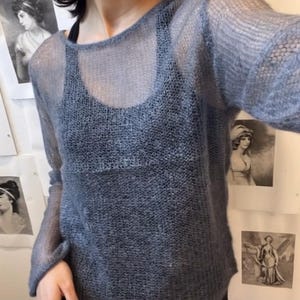 Puede incluir: Un suéter azul claro transparente con mangas largas. El suéter se usa sobre una parte superior negra. La imagen es de una persona que usa el suéter. El fondo incluye obras de arte enmarcadas.