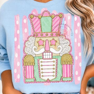 May include: A light blue crewneck sweatshirt featuring a central graphic of a nutcracker in pastel pink, mint green, and gold glitter accents. The nutcracker has star-shaped eyes, a white mustache, and is set against a background of pink brushstrokes.