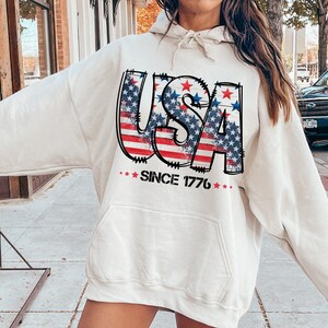 May include: A cream-colored hoodie with a graphic of the word "USA" in red, white, and blue with stars. The hoodie has a drawstring hood and a kangaroo pocket. The text "Since 1776" is printed below the graphic.