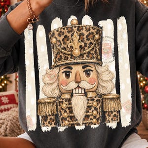 May include: A dark gray sweatshirt featuring a graphic of a nutcracker wearing a leopard print uniform and hat. The nutcracker has a white mustache and beard, and is centered over vertical white brushstroke stripes with white polka dots.