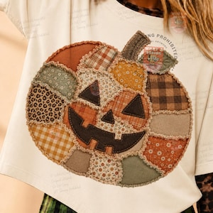 May include: An off-white short-sleeve t-shirt featuring a large pumpkin graphic composed of various fabric-like patches in shades of orange, brown, green, and tan. The pumpkin has a dark brown jack-o'-lantern face and a frayed, textured edge.
