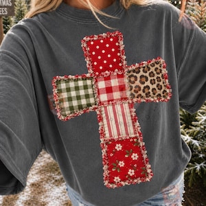May include: A charcoal gray short-sleeve t-shirt featuring a large cross graphic on the front. The cross is composed of five distinct fabric-style patches with red and white polka dots, green and white gingham, red and white plaid, leopard print, and red floral patterns.