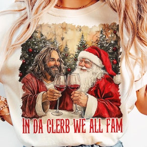 May include: A white t-shirt with a vintage-style illustration of Jesus and Santa Claus toasting with glasses of wine. The text "In Da Clerb We All Fam" is printed below the image.