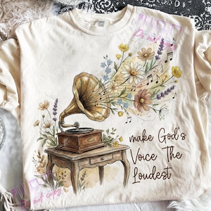 以下が含まれることがあります： クリーム色のTシャツ。ヴィンテージの蓄音機のイラスト、花、音符が描かれています。「make God's Voice The Loudest」の文字入り。柔らかい素材で作られています。