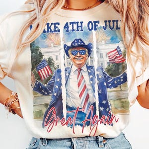 May include: A white t-shirt with a graphic print of a man wearing sunglasses and a cowboy hat, holding American flags. The text "Make 4th of July Great Again" is printed on the shirt.