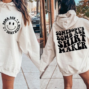 May include: Cream-colored hoodie with a front pocket and drawstring hood. The front features a smiley face design with the text "BOMB ASS SHIRT MAKER". The back of the hoodie displays the text "SOMEBODY'S BOMB ASS SHIRT MAKER" in a retro font.