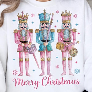 May include: White sweatshirt with a pink, blue, and gold watercolor design of three nutcracker soldiers. The text "Merry Christmas" is printed below the design.