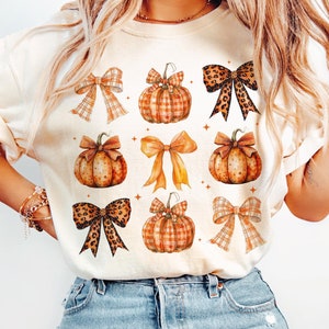 May include: A white t-shirt with a fall-themed design featuring orange and brown pumpkins with plaid and leopard print bows.