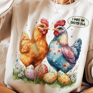 May include: A white sweatshirt with a graphic of two chickens and colorful Easter eggs. The text "I was an Easter egg" is in a speech bubble above the chickens.