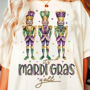 May include: White t-shirt featuring a design of three purple, green and gold Mardi Gras nutcracker figures. The text "it's Mardi Gras y'all" is printed below the figures.