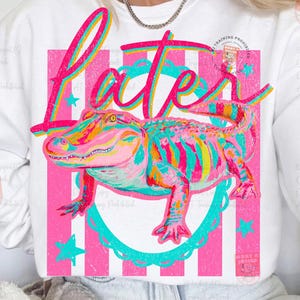 May include: White sweatshirt featuring a colorful alligator design with the word "Later" in a vibrant, layered font. The design includes pink stripes, teal accents, and stars, creating a playful and eye-catching graphic.