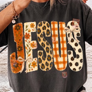Könnte beinhalten: Ein anthrazitfarbenes T-Shirt mit dem Wort JESUS in großen Buchstaben. Jeder Buchstabe hat ein einzigartiges Muster: orangefarbene Blumen, cremefarbene Blumen, Leopardenmuster, orangefarbenes Vichy-Karo und cremefarben mit schwarzen Punkten, verziert mit zwei beigen Schleifen.